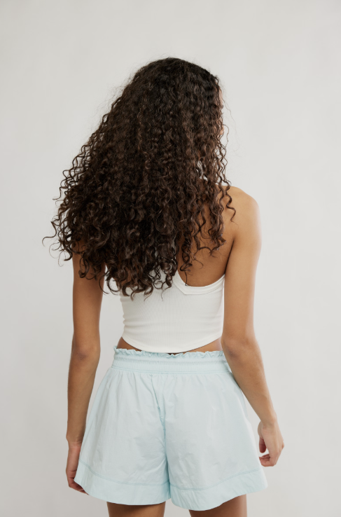 A woman with long, curly dark brown hair is shown from behind, wearing a white ribbed tank top and light blue shorts against a plain light gray studio background.