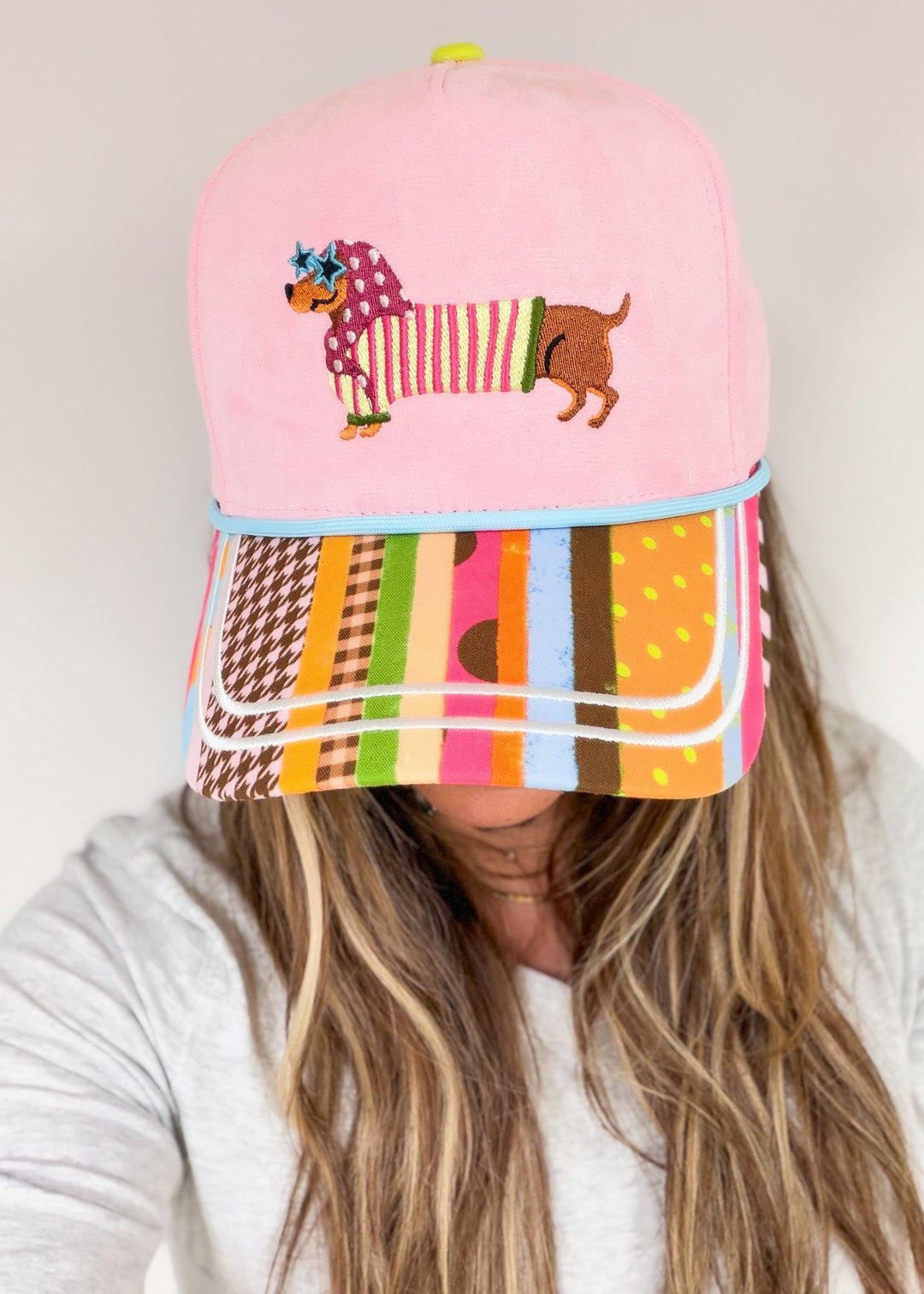 Woman wearing pink faux suede hat with doxie on front wearing bandana and sunglasses with striped bill on plain background