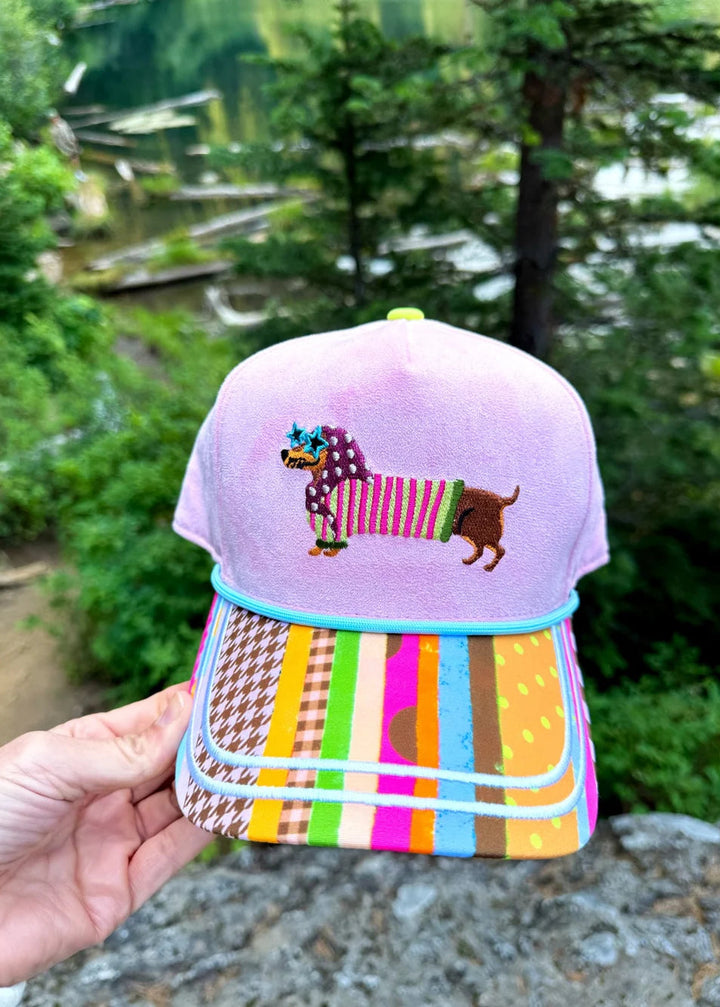 Pink faux suede trucker hat with doxie on front wearing bandana and sunglasses with stripe bill outdoors