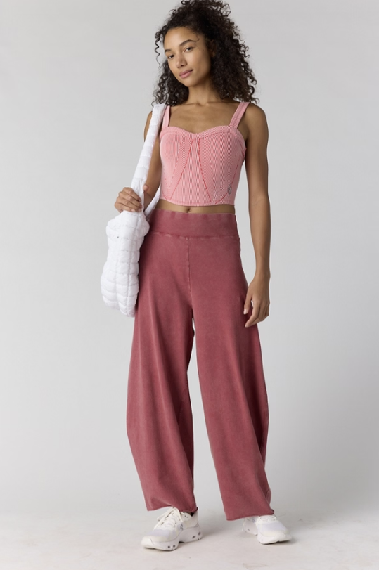 A woman modeling a two-piece outfit consisting of a light pink knit crop top and dusty rose wide-leg pants, styled with white sneakers and a white quilted shoulder bag against a light gray studio background.