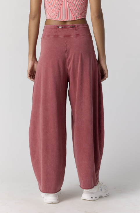 A person shown from the waist down wearing loose, wide-leg dusty rose pants with a textured fabric, paired with a pink ribbed crop top and white sneakers, against a light gray studio background.