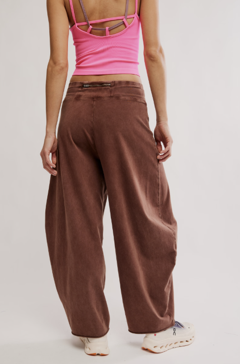 A person shown from the back wearing wide-leg brown pants, a bright pink strappy crop top, and white sneakers against a plain light gray studio background.