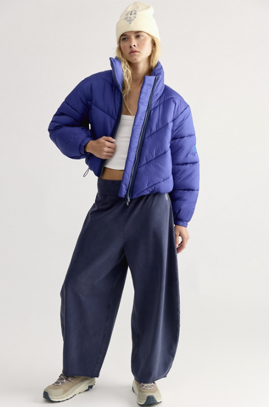 A model wearing a vibrant blue cropped puffer jacket, navy wide-leg pants, a cream beanie, and beige sneakers, standing against a light gray studio background.