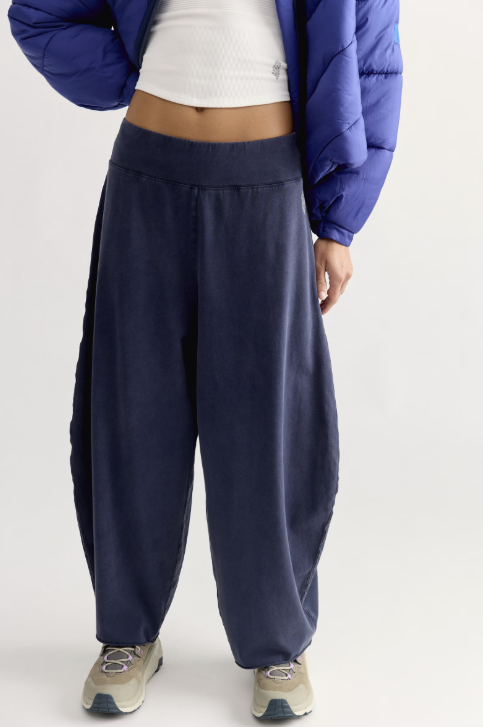 A person wearing loose navy wide-leg sweatpants, styled with a white ribbed crop top, a blue puffer jacket, and beige sneakers, photographed against a light gray studio background.