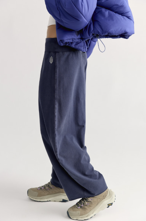 A person shown from the waist down wearing loose navy blue sweatpants, beige hiking shoes, and holding a blue puffer jacket against a plain light gray studio background.