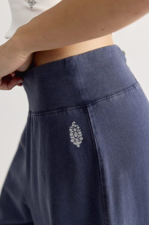 Close-up of a person wearing navy blue high-waisted lounge pants, pulling the waistband slightly to show stretch, with a small white embroidered pinecone logo on the front.