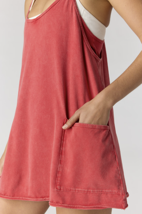 A close-up of a model wearing a washed coral-red sleeveless tank dress with a large front patch pocket, layered over a white tank top, against a light gray studio background.