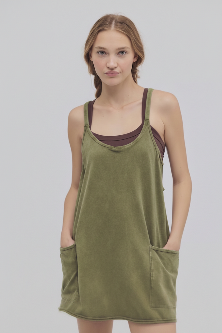 A woman modeling an olive green sleeveless slip dress with front patch pockets over a dark plum tank top, shot against a light gray studio background.
