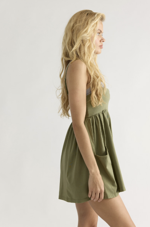 A blonde woman shown in side profile wearing a sleeveless olive green mini dress with a gathered waist and side pocket, photographed against a plain light gray studio background.