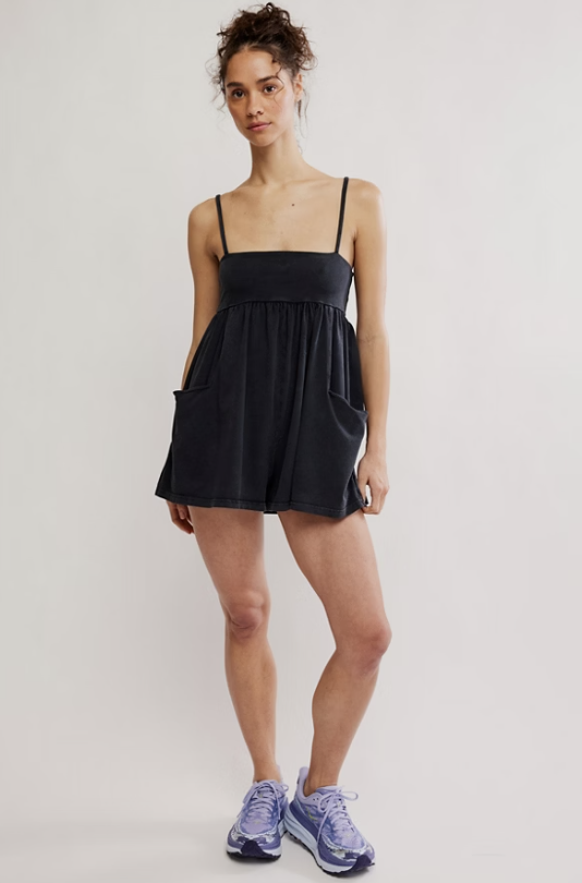 A woman modeling a black sleeveless romper with thin shoulder straps, a gathered waist, and side pockets, paired with light purple sneakers, against a plain light gray studio background.