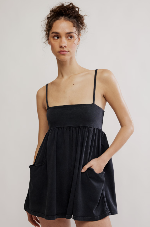 A woman modeling a black spaghetti-strap mini dress with a square neckline, gathered waist, and side pockets against a plain light gray studio background.