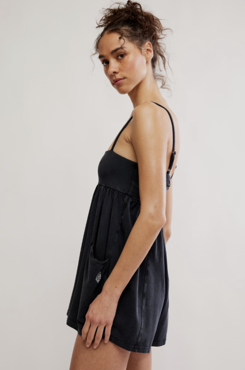 A woman modeling a black sleeveless mini dress with thin straps and a gathered skirt, photographed against a plain light gray studio background.