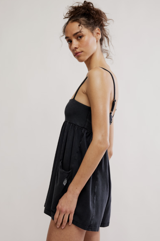 A woman modeling a black sleeveless romper dress with thin straps and a gathered waist, photographed against a plain light gray studio background.