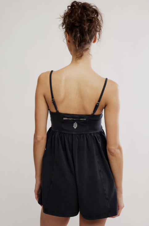A woman viewed from behind wearing a black camisole-style romper with thin straps, a gathered waistband, and short flared legs against a plain light background.