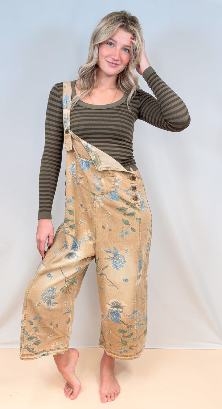 Woman wearing beige floral overalls with green stripe long sleeve shirt on plain background