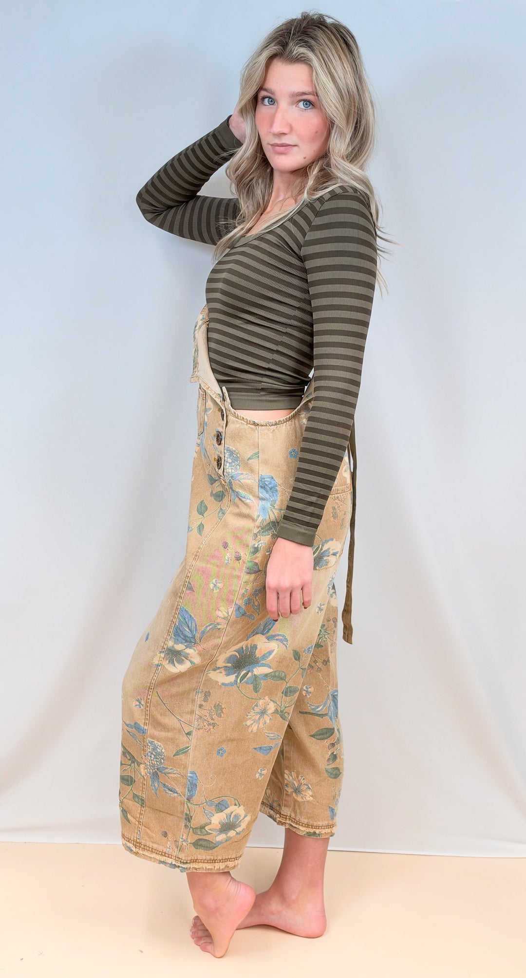 Woman shown from side view wearing beige floral overalls with green stripe long sleeve on plain background