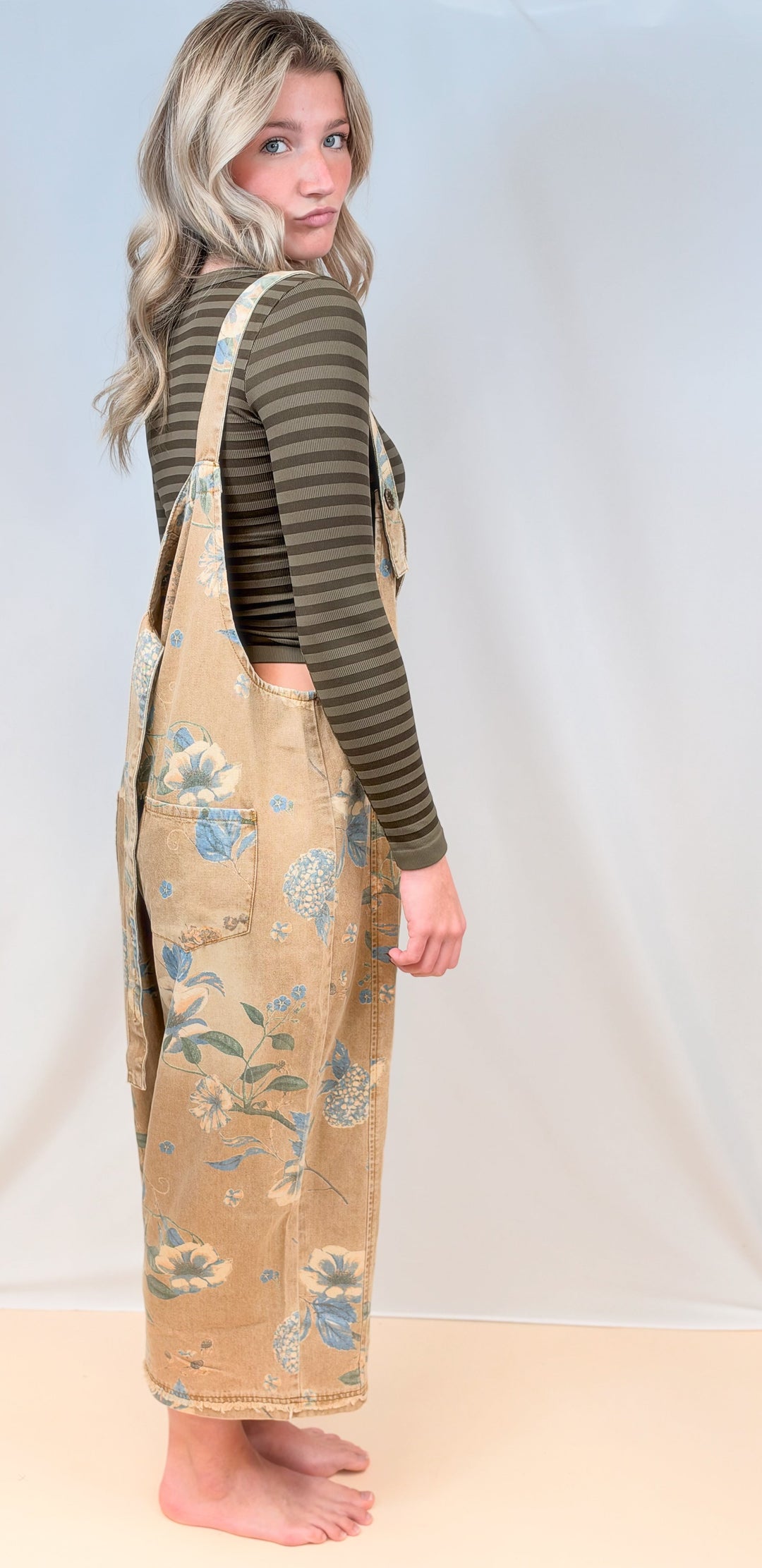 Woman shown from side view wearing beige floral overalls and green stripe long sleeve top on plain background