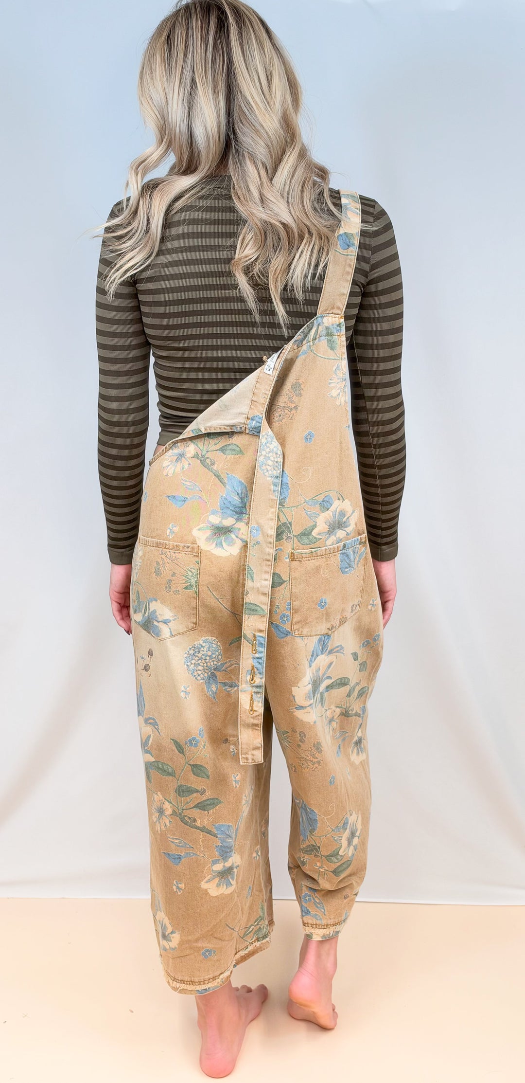 Woman shown from back view wearing beige floral overalls with green stripe long sleeve top on plain background