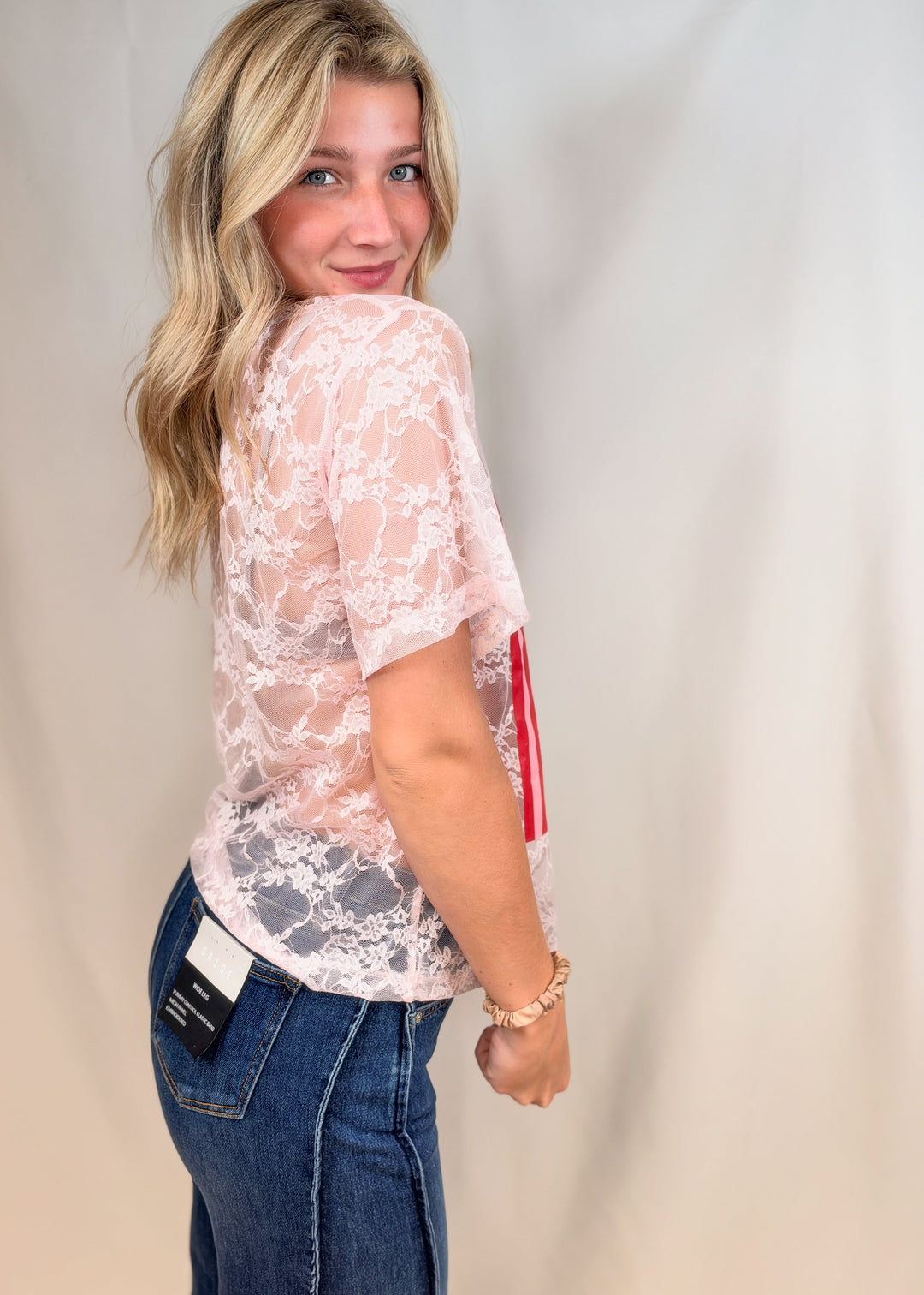 Woman shown from side view wearing pink lace tee and denim jeans on plain background