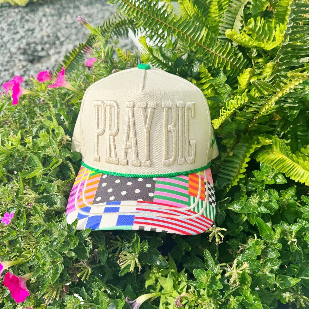 Cap with 'PRAY BIG' text on a colorful band, placed on green foliage.