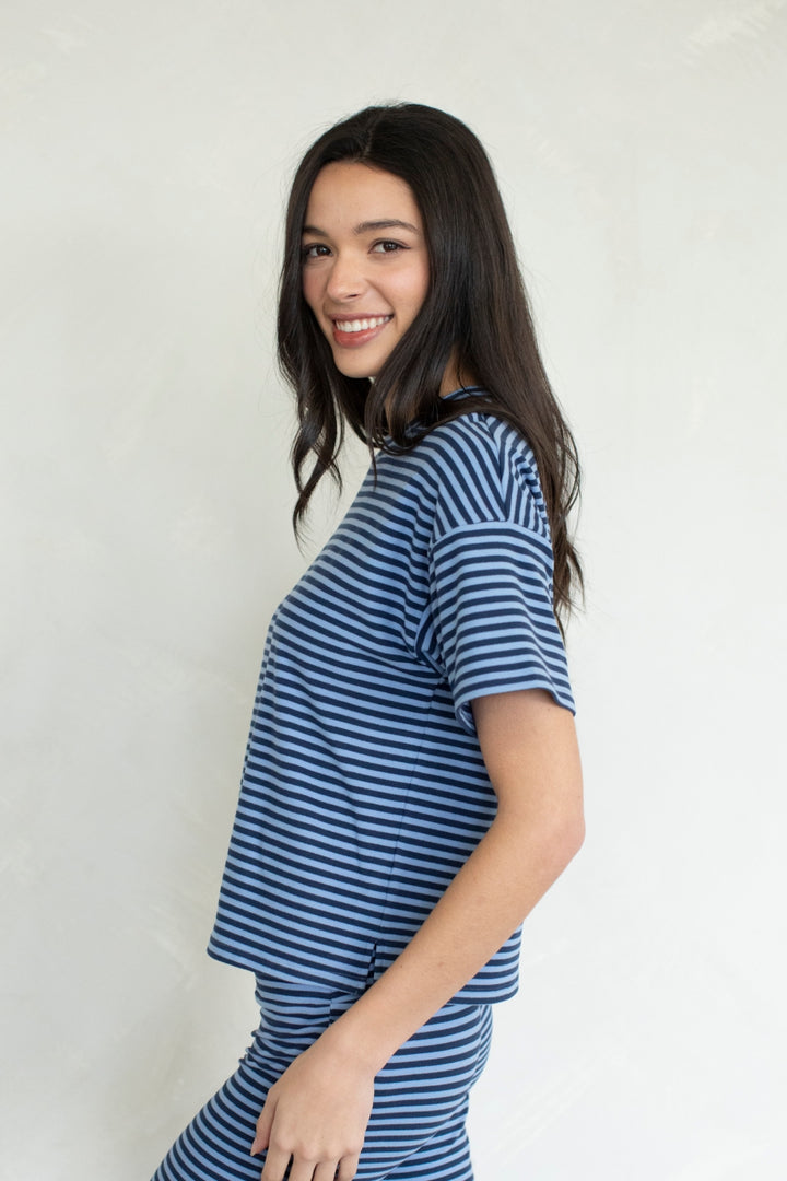 A woman with long dark hair is shown from the waist up, wearing a blue and white horizontally striped short-sleeve top and matching striped bottoms, posing against a plain white wall.