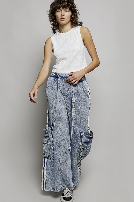 A model wearing a white sleeveless top and wide-leg, acid-wash blue denim cargo pants with side stripes, standing against a neutral gray studio background.