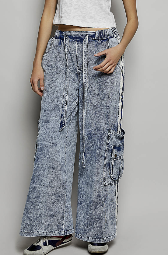A person wearing wide-leg acid-wash denim cargo pants with side pockets and a drawstring waist, paired with a white cropped top and sneakers, against a plain light gray studio background.