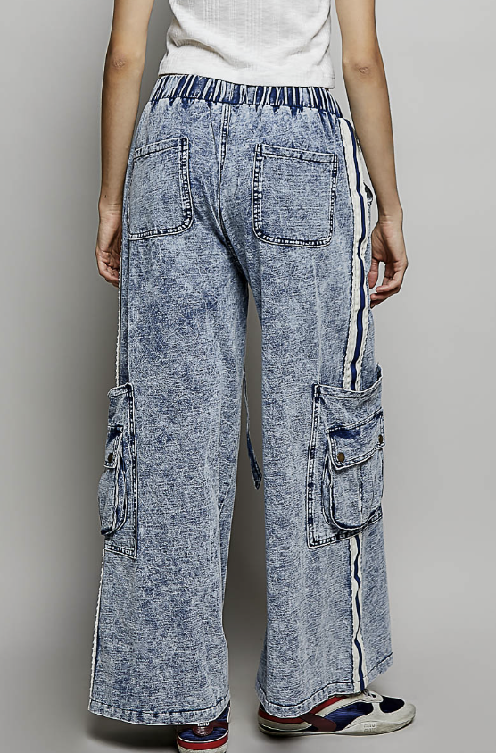 A person shown from the back wearing wide-leg acid-wash denim cargo pants with side pockets and a striped elastic waistband, paired with a white top and sneakers against a light gray studio background.