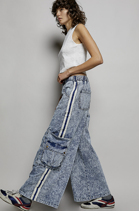 A model wearing wide-leg, light-wash blue denim cargo pants with vertical white and navy stripes down the sides, paired with a white tank top and sneakers, set against a plain gray studio background.
