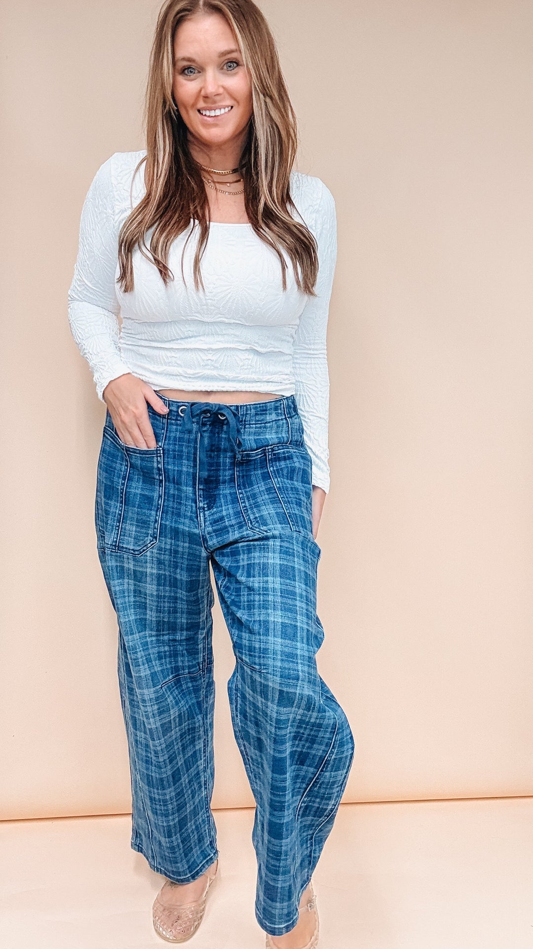 A woman modeling a white ribbed long-sleeve crop top and blue plaid wide-leg pants against a solid beige studio backdrop.