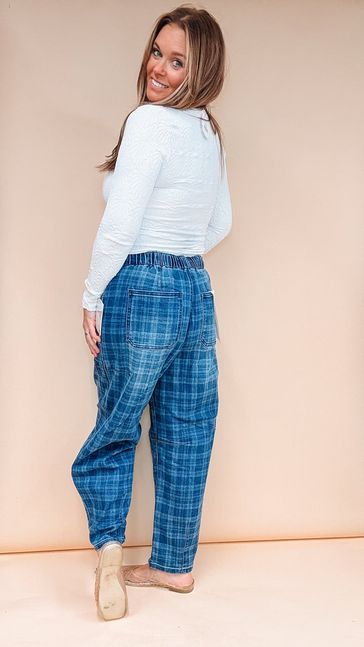 A woman wearing blue plaid lounge pants and a white long-sleeve top, photographed from the back against a solid beige studio backdrop.