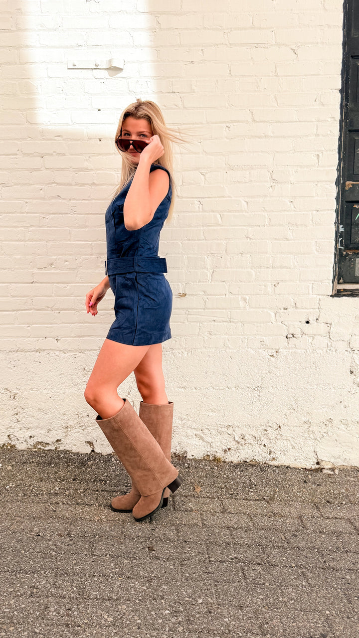 Woman wearing suede navy mini dress with tall beige boots on white brick background