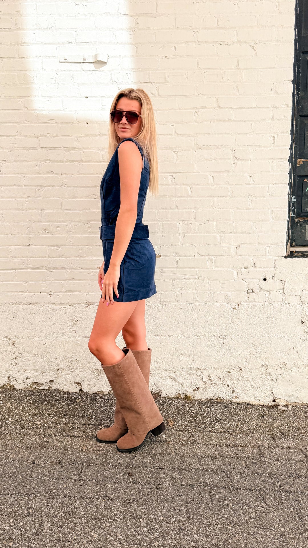 Woman wearing navy suede mini dress with tall beige boots on white brick background