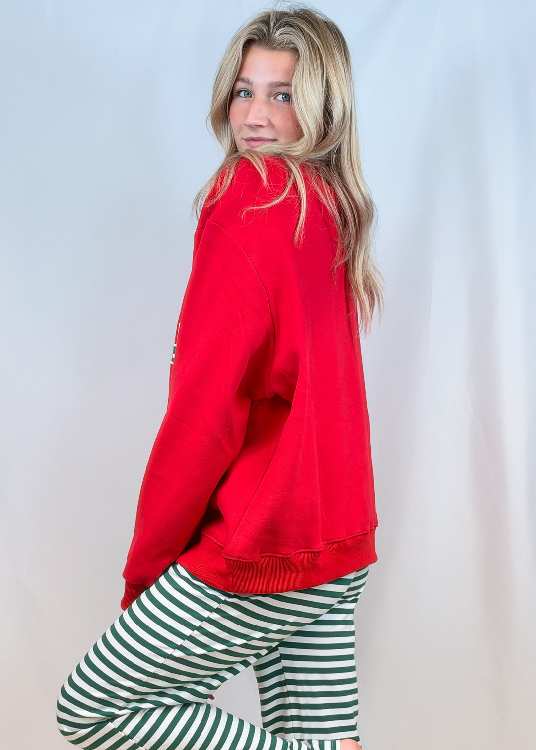 Woman shown from side view wearing red sweatshirt with green and white stripe pants on plain background