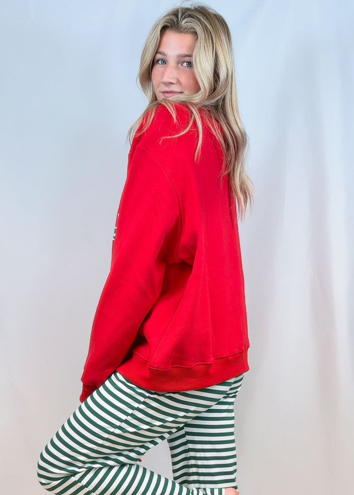 Woman shown from side view wearing red sweatshirt with green and white stripe pants on plain background