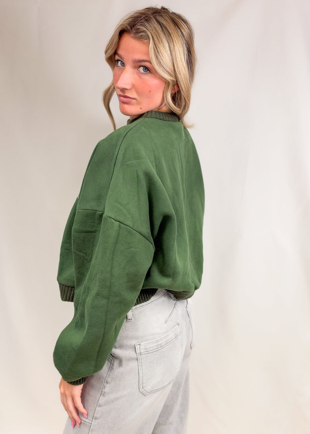 Woman wearing green mock neck sweatshirt and grey jeans on plain background