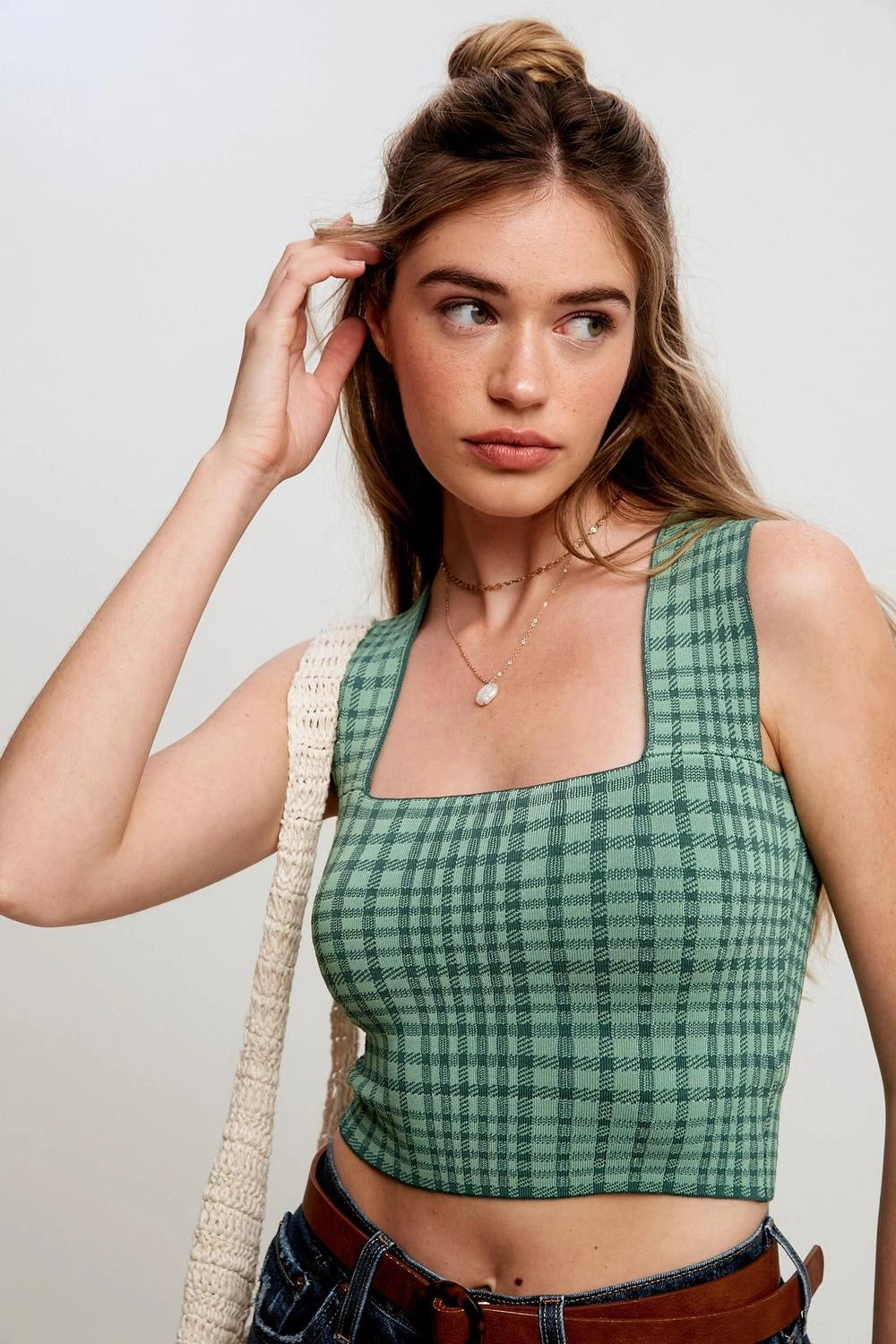 A woman wearing a green and black plaid square-neck crop top, paired with a cream woven shoulder bag and blue jeans, photographed against a light gray studio background.