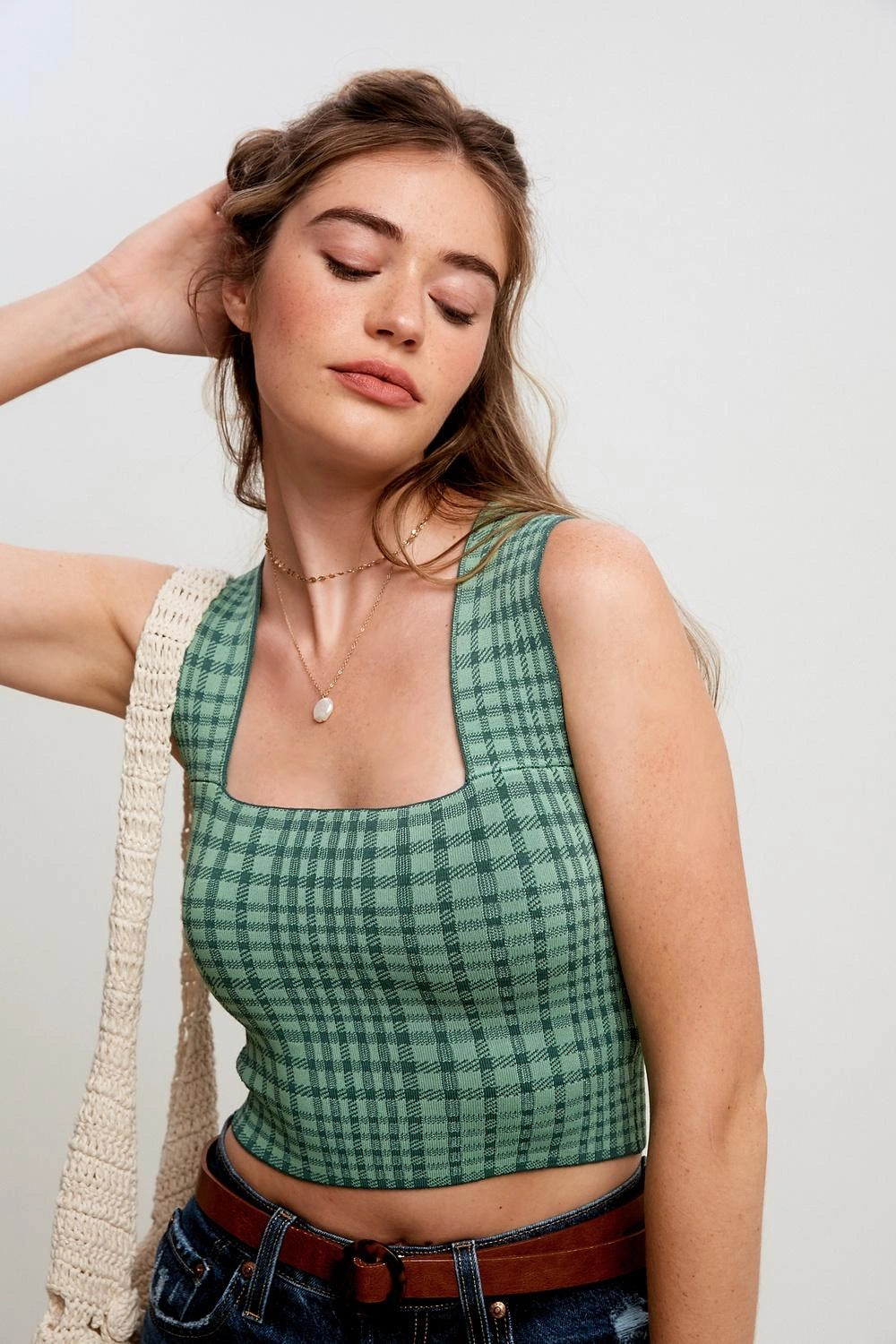 A woman wearing a green and black plaid sleeveless crop top, styled with a cream crochet shoulder bag, a brown belt, and blue jeans against a plain light gray background.