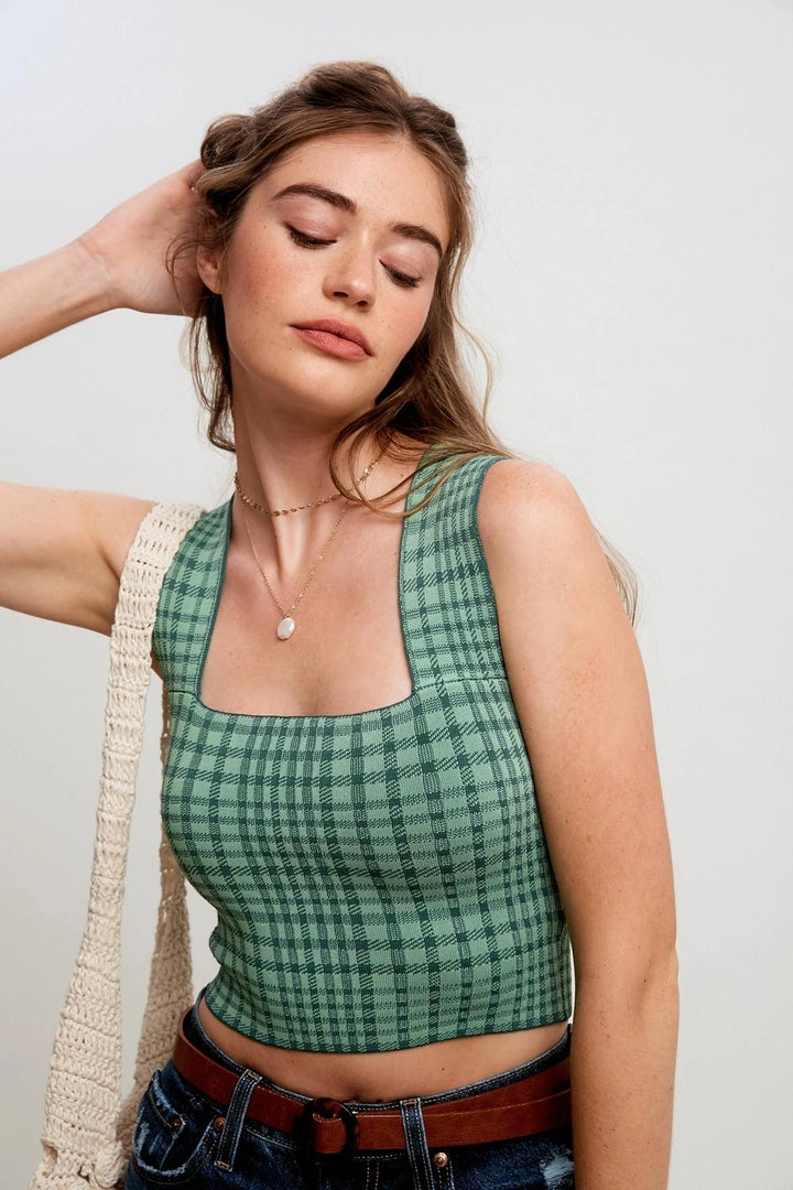 A woman wearing a green and black plaid sleeveless crop top, styled with a cream crochet shoulder bag, a brown belt, and blue jeans against a plain light gray background.