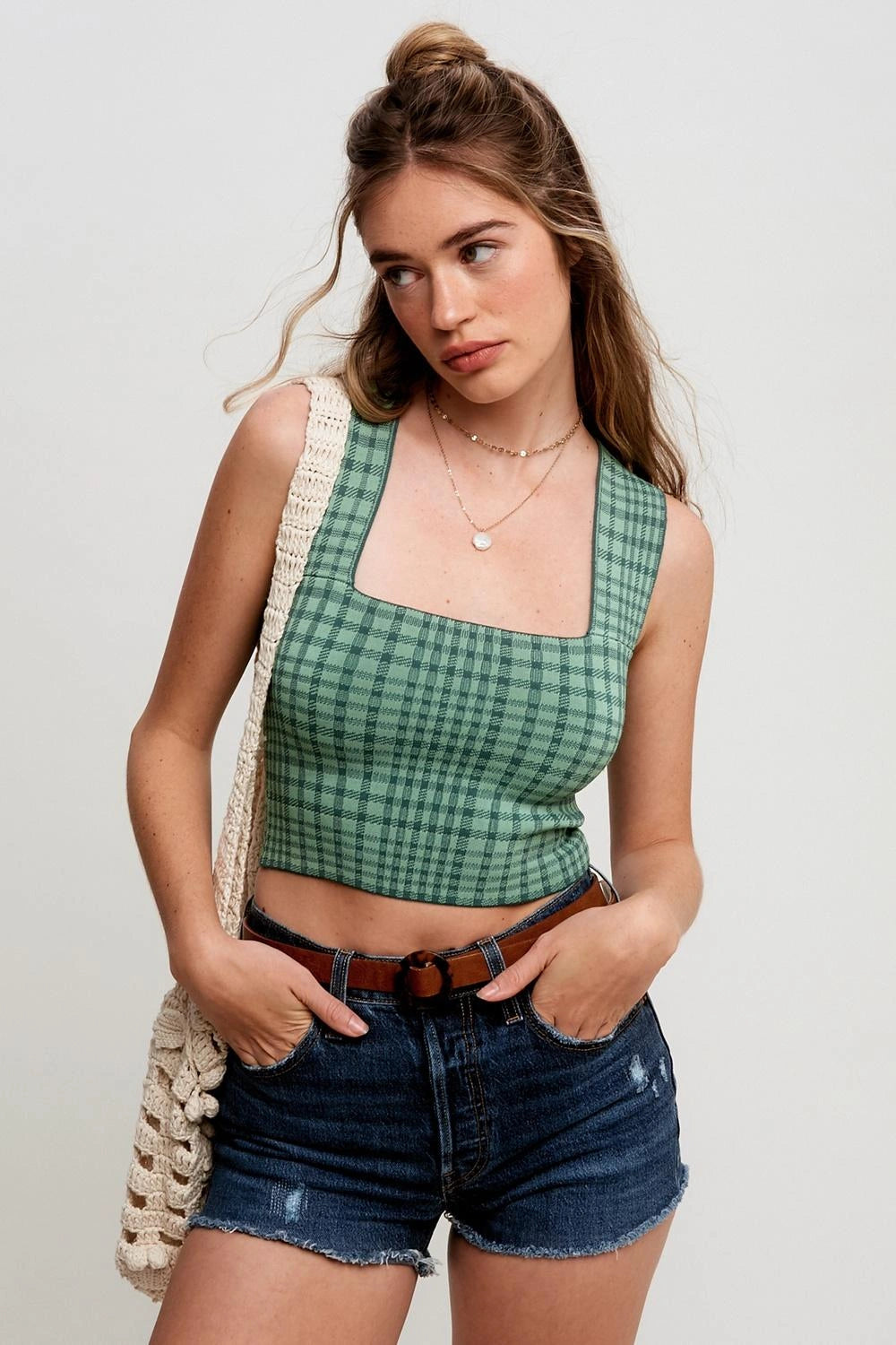 A young woman wearing a green and blue plaid crop top with a square neckline, paired with distressed denim shorts and a cream woven shoulder bag, posed against a light gray studio background.