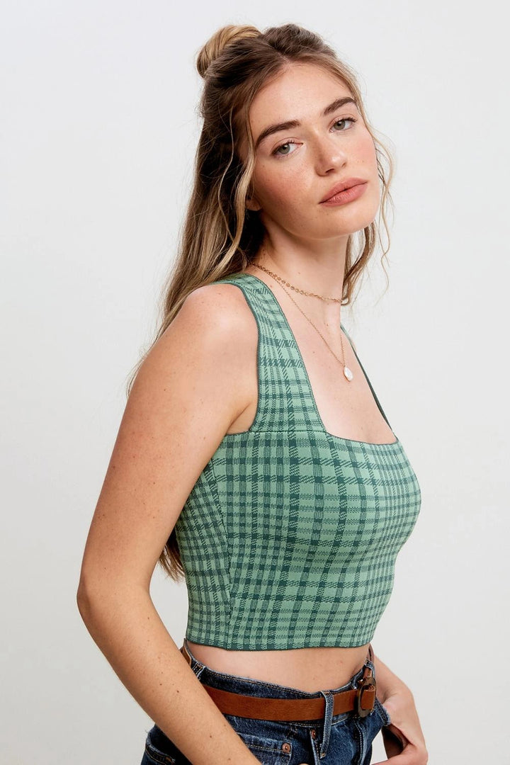A woman wearing a green and black checkered crop top with a square neckline, paired with blue jeans and a brown belt, photographed against a light gray studio background.