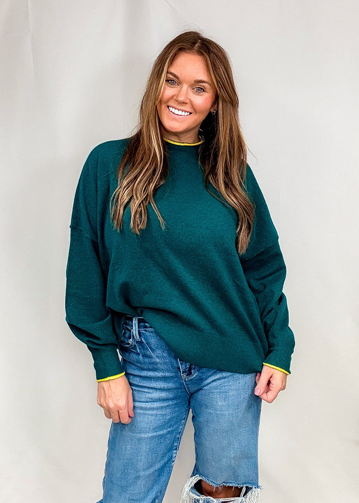 Woman wearing dark green sweater with bright trim on neck and sleeves with jeans on plain background