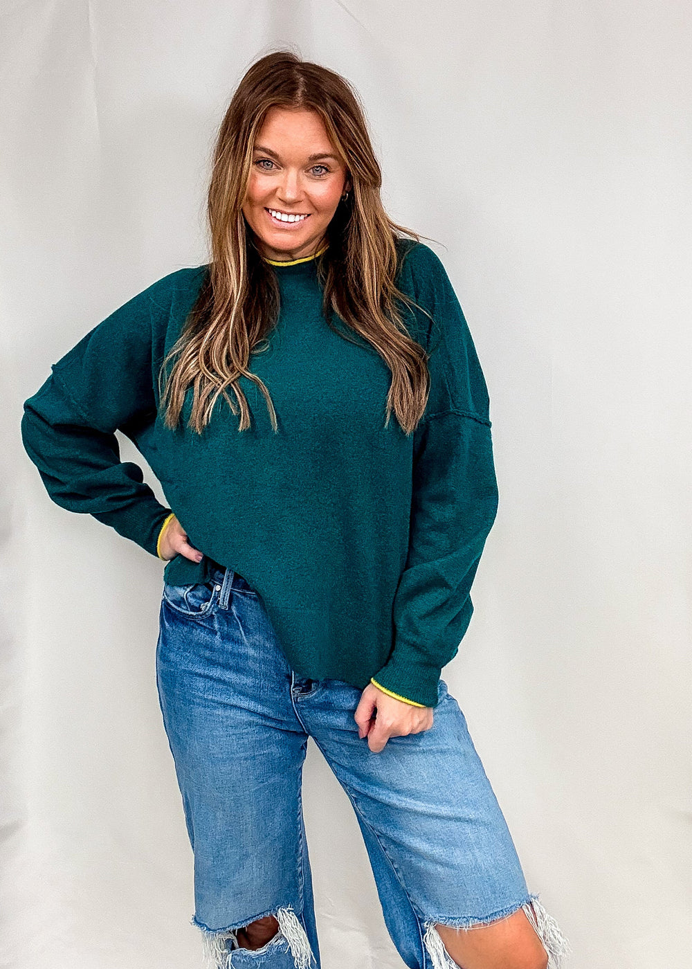 Woman wearing dark green sweater with bright trim on neck and sleeves with jeans on plain background