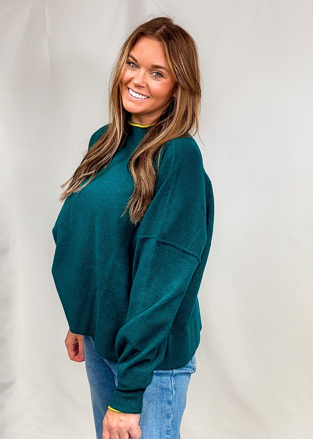 Woman wearing dark green sweater with bright trim on neck and sleeves with jeans on plain background