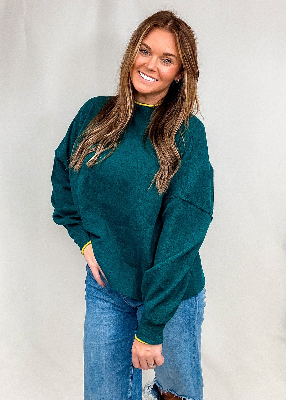 Woman wearing dark green sweater with bright trim on neck and sleeves with jeans on plain background