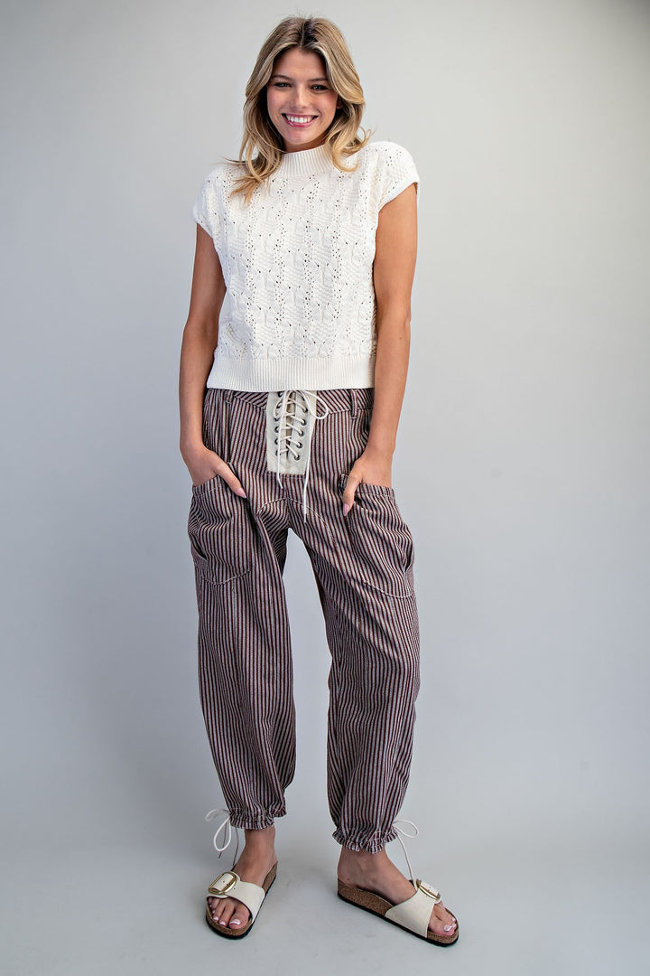 A woman modeling a white textured sleeveless top and brown and white vertically striped drawstring pants, paired with beige slide sandals, against a light gray studio background.