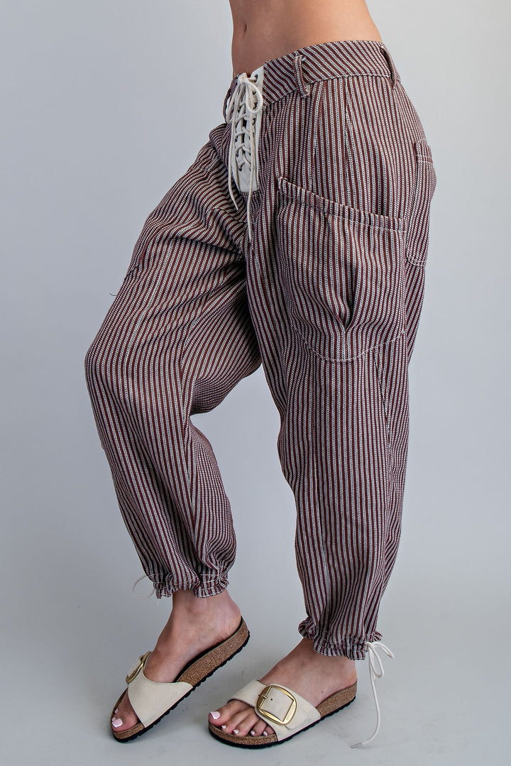 A person wearing loose brown and white vertically striped drawstring pants with large patch pockets and cuffed ankles, paired with cream slide sandals, against a light gray studio background.