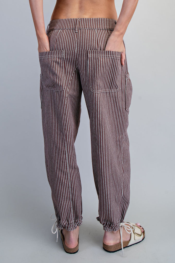 Rear view of a person wearing loose, vertically striped pants in muted brown and white, with hands in the back pockets and sandals, against a light gray studio background.