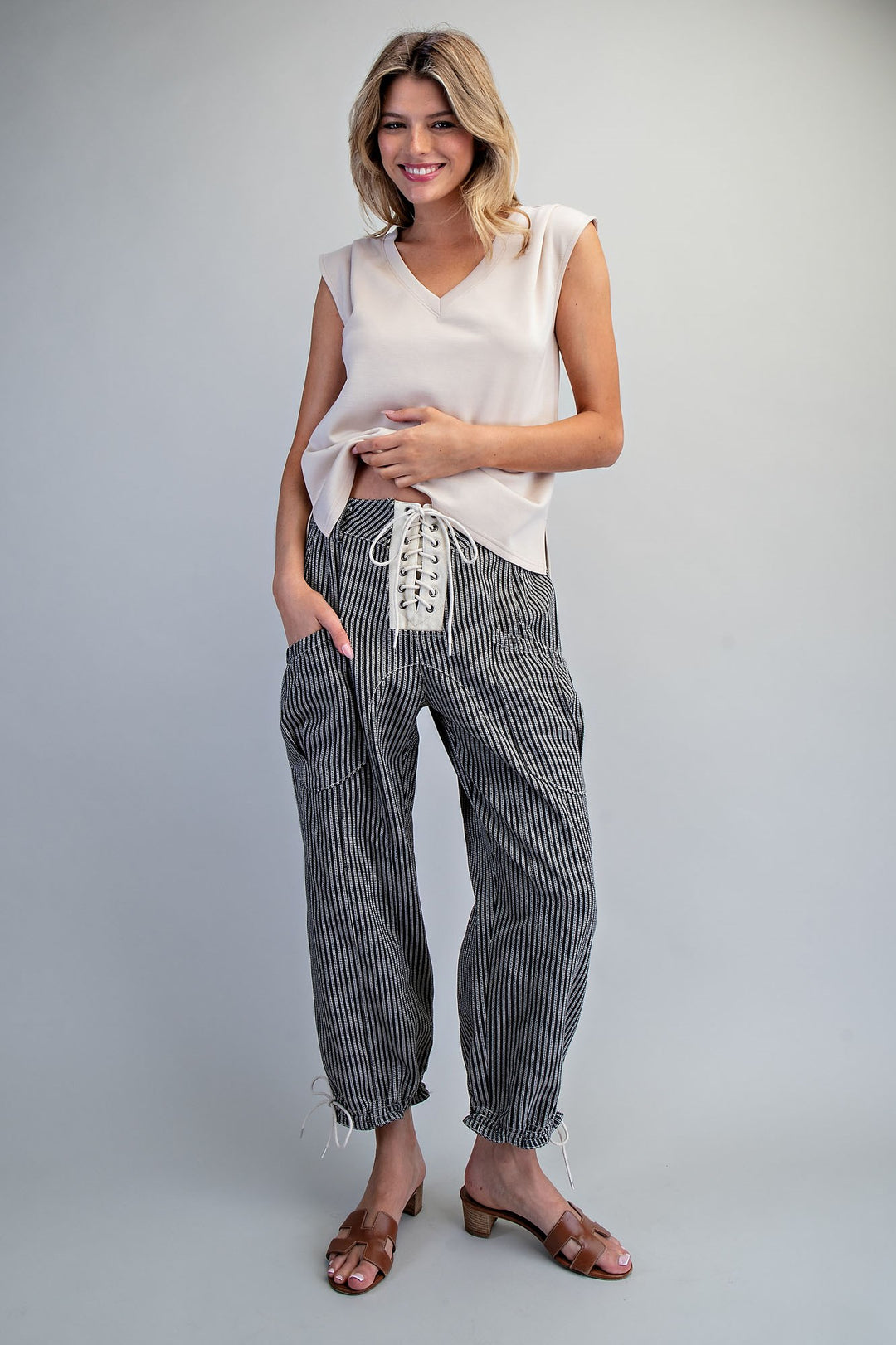 A woman modeling a sleeveless cream V-neck top and black-and-white vertically striped cropped pants with lace-up detailing at the waist, paired with brown heeled sandals, against a light gray studio background.