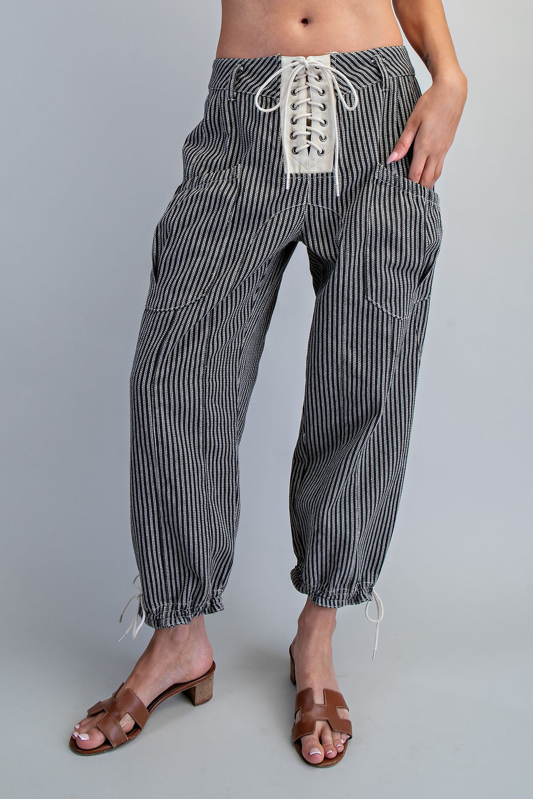 A person wearing loose black-and-white vertically striped pants with a lace-up front waist, shown from the waist down against a light gray studio background.